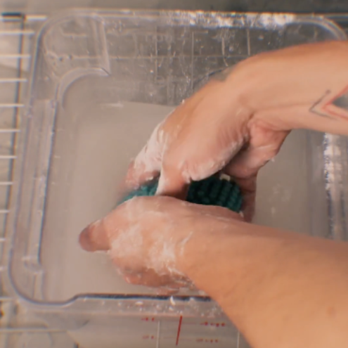 A close-up of a person's hands covered in white powder, scrubbing a dark green textured item inside a clear plastic tub filled with cloudy liquid.