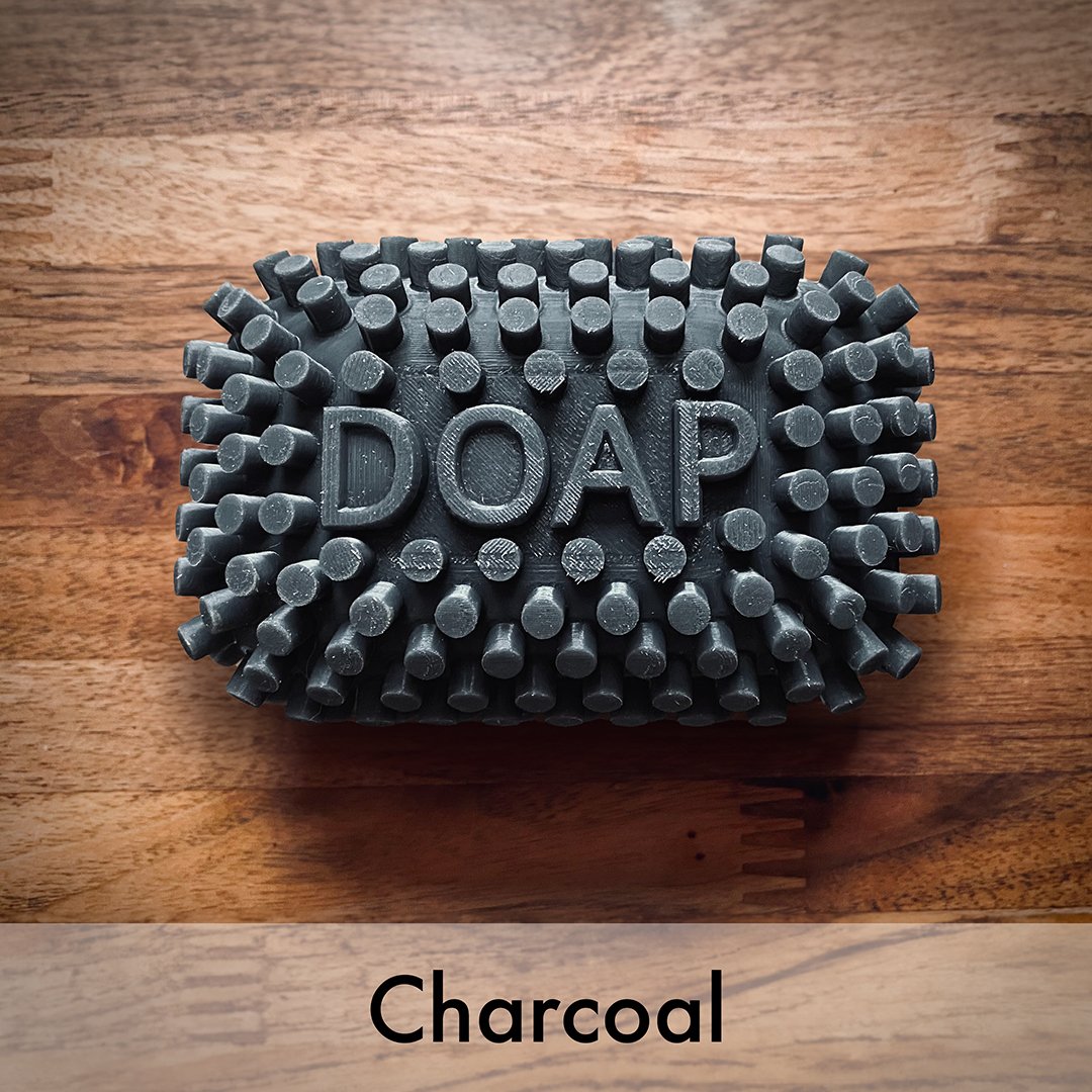 A very dark grey textured charcoal DOAP bar embossed with the word 'DOAP' on a wooden surface, with the word 'Charcoal' printed below.