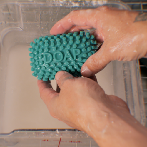 A close-up of hands holding a teal green textured silicone bar embossed with the word 'DOAP' over a clear plastic tub of flour water.