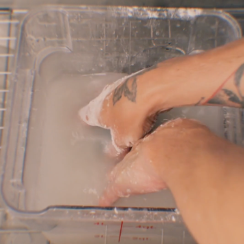 A close-up of bakers hands submerged in a clear plastic tub filled with cloudy liquid, about to use a DOAP bar to clean sticky dough from the bakers hands.