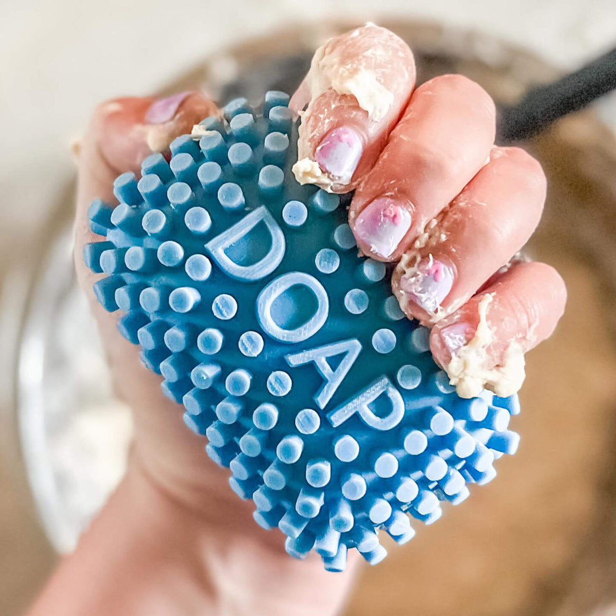 A close-up of a hand holding a blue textured scrubber labeled 'DOAP' with sticky dough on the fingers, set against a blurred background of a bowl and countertop.