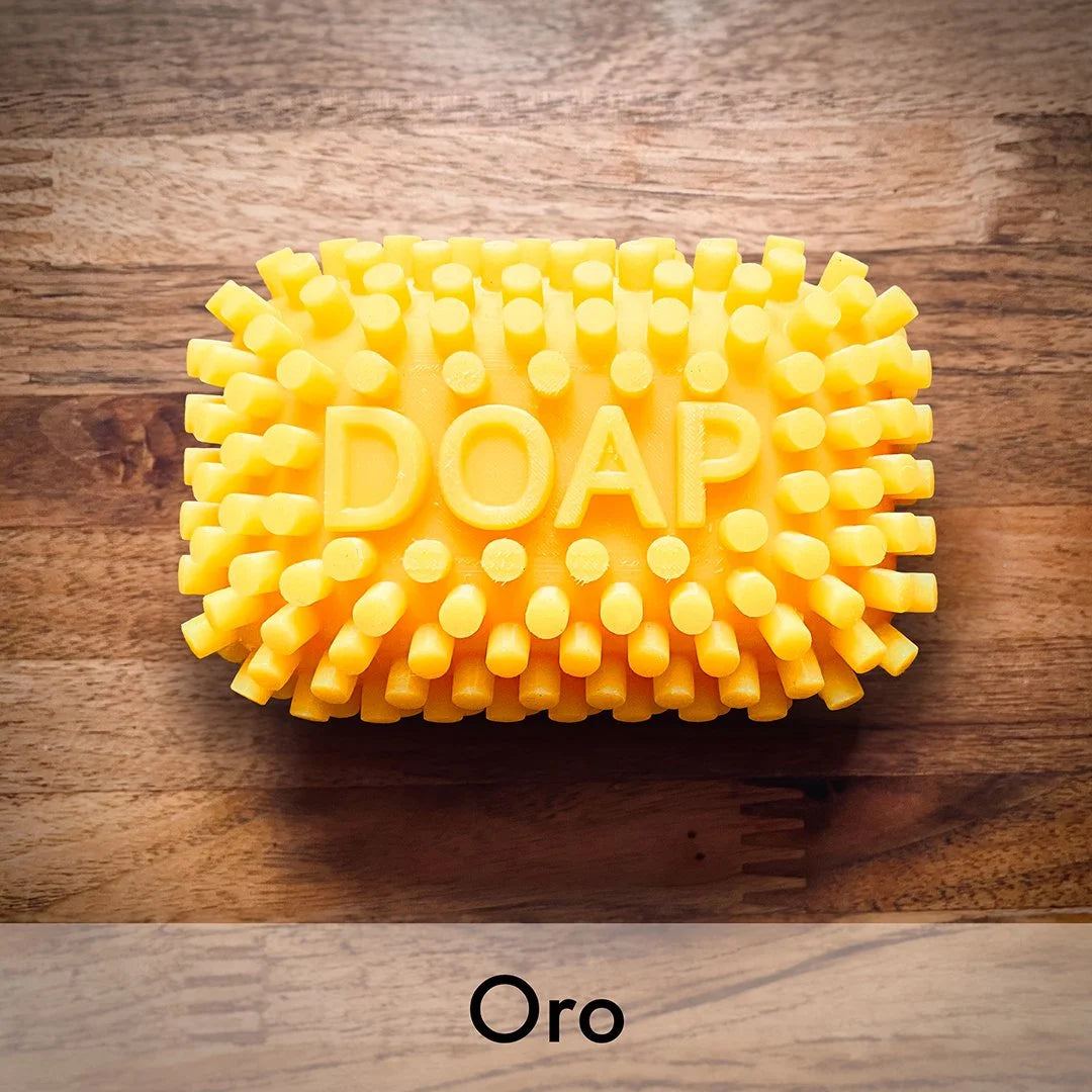 A yellow textured soap bar with the word 'DOAP' embossed in the center, placed on a wooden surface. The image has a wood grain background and a bottom banner with the text 'Oro'.
