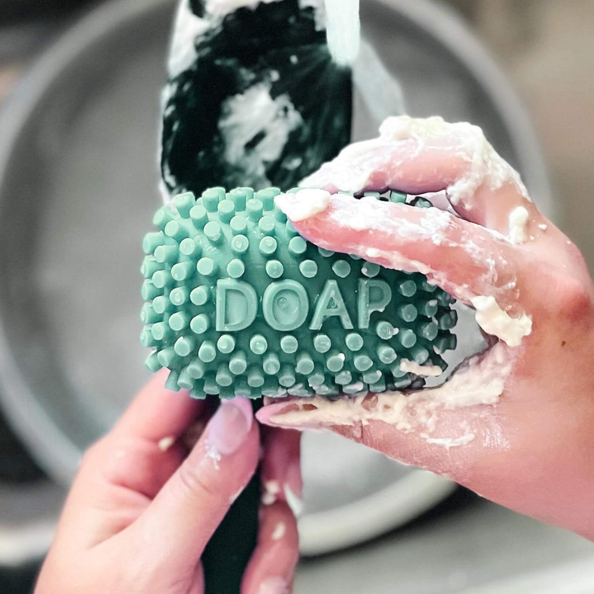 A close-up of a person washing sticky dough off a spatula with a green textured  silicone bar labeled 'DOAP' under running water in a sink. The hands are covered in sticky dough, and the background shows a blurred sink basin.